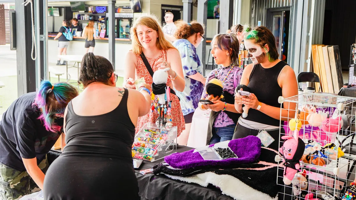 Attendees browse a vendor table of handmade jewelry, keychains and plushies while a vendor helps a smiling shopper, with a concession stand in the background at Knox Pop Con.